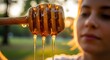 © Richafuji - Close up of honey dripping from a wooden dipper with a blurred woman and green background. Warm lighting highlights the amber liquid and the texture of the wood.