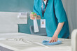 © AnnaStills - Middle aged Caucasian woman cleaning hospital bed with spray bottle and cloth, wearing medical uniform and identification badge, performing sanitation in healthcare facility