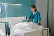 © AnnaStills - Young adult Caucasian woman nurse making hospital bed in patient room, standing beside medical equipment, wearing identification badge, preparing clean linens for next patient