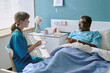 © AnnaStills - Caucasian young adult female nurse sitting beside hospital bed talking to middle aged biracial man patient lying under blanket holding hands during medical consultation