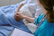 © AnnaStills - Senior Caucasian patient lying in hospital bed receiving comforting touch from young adult Caucasian female nurse, medical form and stethoscope visible in clinical setting