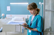© AnnaStills - Young adult Caucasian woman nurse standing in hospital room using digital tablet, focusing on patient records, medical professional working in healthcare environment