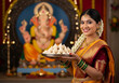 © Stockphotosbysumit - Portrait of a beautiful traditional Indian woman in a silk saree, holding a plate of modak prasad as an offering during the Ganesh Chaturthi festival, with a Ganesha idol in the background.