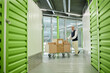 © Seventyfour - Middle aged Caucasian man pushing cart loaded with cardboard boxes through corridor of self storage facility, looking focused while transporting belongings in modern warehouse environment