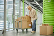 © Seventyfour - Middle aged Caucasian woman writing on cardboard box while standing next to cart loaded with packages in modern storage facility, handling logistics and inventory management tasks