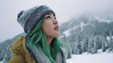 A young woman with dyed green hair looks up thoughtfully, wearing a winter hat and scarf against a backdrop of snow-covered mountains and trees on a cold day.