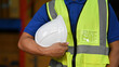 © saltdium - Industrial safety concept, Close-up of a warehouse worker in a safety vest holding a white hard hat, symbolizing workplace safety and protection