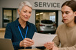 © Jens - Consultation at the Service Center: An older female service advisor discusses a document with a young customer in the service bay of a vehicle repair facility.