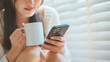 © oatawa - Closeup of a young Asian woman sitting by the window, holding a warm drink while checking messages on her mobile phone – cozy lifestyle, peaceful indoor moment, digital comfort and modern relaxation