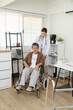 © Ekkasit A Siam - Orthopedic doctor prepares to assist elderly female patient in wheelchair during physical therapy session for joint pain, medical setting with walking aid and treatment table in clinic room