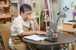 © Ekkasit A Siam - Asian elderly female florist sitting alone looking stressed while doing account review in flower shop using laptop and paper notebook reflecting challenges of small business management