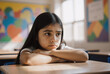 © Running opossum - Thoughtful young Latina girl sitting alone in a colorful classroom during sunny afternoon with contemplative expression