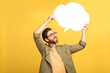 © Prostock-studio - A middle aged man in casual attire stands against a vibrant yellow backdrop, smiling and holding a blank speech bubble above his head as if about to share a thought or message.