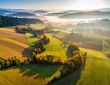 Widok z góry na spowite w lekkiej mgle zielone pola uprawne, łąki i drzewa o wschodzie słońca
