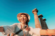 © EdNurg - Asian tourist with hat taking a selfie with outstretched arms in front of ancient greek columns with a beautiful blue sky