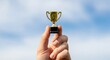 © Pixel Stone - Tiny Golden Trophy Held in Hand Against Blue Sky Background