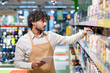 © Liubomir - An employee in a beige apron checks products on a supermarket shelf while holding a tablet.