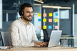 © Liubomir - A smiling man wearing headphones uses a laptop in a modern office setting. He has glasses and is working on a project.