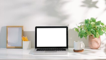 Wall Mural - Laptop with blank white screen on a white desk, accompanied by coffee mug, and green plant in natural light.