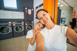 © dtatiana - Mother and daughter taking a selfie while waiting in a self-service laundromat, looking at camera and bonding during laundry time, promoting shared responsibilities and everyday routine