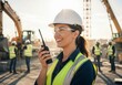 © tebu - Smiling woman in hardhat and safety vest holding a walkietalkie on a construction site