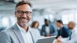 © Formoney - Smiling man in formal attire holds a tablet in a modern office setting. Behind him, colleagues engage in a discussion, creating a collaborative vibe during work hours.