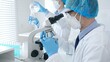 © rogerphoto - Masked scientist examining specimen under microscope while laboratory assistant handles glassware in a professional medical investigation. Health care and science concept