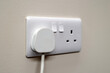 © Tosh Lubek - White UK 3-pin mains plug inserted into modern dual wall socket with switches. Mounted on a light painted wall. Suitable for illustrating household electricity, safety, and electrical infrastructure