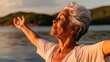 © buraratn - Senior woman enjoys tranquility of lake, stretching her arms wide in moment of peace and connection with nature. serene landscape and warm sunlight enhance her joyful expression