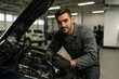 © Denny - A young male auto mechanic with light beard holds a wrench by an open car hood, focused on repairing the engine.