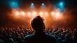 © Dulemegapixel - A silhouette of a performer stands before a vibrant crowd, illuminated by bright stage lights that create an atmosphere of excitement and energy during a live performance.