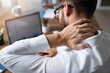© David Zarzosa - Male worker with neck pain working at desk with laptop in office environment symbolizing stress and overwork.