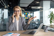 © Liubomir - A smiling businesswoman in a grey suit is talking on the phone while gesturing with her hand, working from the office.