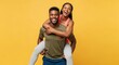 © mohammed - A joyful African American couple in activewear having fun. The strong man gives his happy girlfriend a piggyback ride on a yellow background.