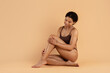 © Anastasiya - Smiling black woman in underwear sitting cross legged on beige background and looking down in a soft, peaceful pose. Warm tones and calm posture for beauty, body care, and inner balance visuals