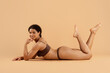 © Anastasiya - Young African American woman in underwear lying on stomach, smiling and looking at camera while posing on beige background. Ideal for wellness, body confidence, and positive lifestyle content