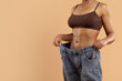 © Anastasiya - Cropped image of toned woman in brown underwear holding oversized jeans. Symbol of significant weight loss, fitness goal achievement, and healthy lifestyle transformation, copy space
