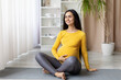 © Anastasiya - Smiling pregnant woman sitting cross legged on yoga mat, gently holding belly. Peaceful moment of prenatal bonding and emotional wellbeing, copy space