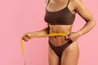 © Anastasiya - Closeup of black woman measuring her waist with a tape while standing on pink background. Fitness and weight management concept focusing on discipline, shape, health goals, and body confidence