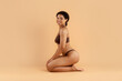 © Anastasiya - Side view of smiling woman sitting on knees in brown underwear against beige background. Soft lighting and confident pose for wellness, beauty, and comfort concepts.