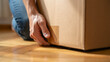 © Lisa - Close-up of a man's hand lifting a cardboard box from a parquet floor in natural light. The moment captures a move or unpacking process