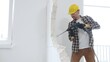 © volha_r - Male construction worker wearing beige checkered shirt, yellow hard hat and protective gloves, is demolishing white wall with rotary hammer drill, generating dust, medium long shot. Renovation project