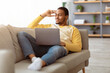 © Prostock-studio - Dreamy black guy with modern laptop resting on sofa at home, looking at copy space. Happy african american young man using notebook and thinking about great future, living room interior
