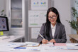 © crizzystudio - Asian businesswoman analyzing financial charts and taking notes in modern office