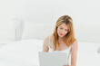 © wavebreak3 - Woman sitting on bed using silver laptop beside white table lamp in minimalist bedroom, copy space