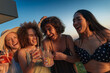 © bernardbodo - Four cheerful young women laughing and drinking cocktails at sunset