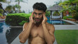 © Krakenimages.com - Shirtless man presses temples at resort pool during sunlit morning break; headache discomfort strain tension.