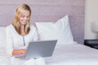 © WavebreakMediaMicro - Middle-aged woman sitting cross-legged on bed typing on laptop in bedroom with pillow and lamp