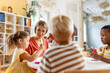 © lordn - Preschool teacher engaging with children at a table during an arts and crafts activity in a bright classroom