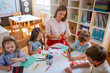 © lordn - Preschool teacher guiding children from diverse backgrounds during a colorful arts and crafts activity in a bright classroom.